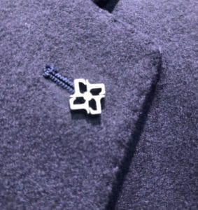 タリアトーレ(TAGLIATORE)のアイコンである、ブランドの頭文字「T」を4つ組み合わせたシルバーのラペルピンのアップ写真。ネイビーの起毛生地(ウール素材)のジャケット襟元のフラワーホールに装着されている様子。