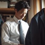 「忙しい朝、普通のクローゼットの前で、ハンガーにかかったネイビーのスーツジャケットを手に取り、複雑な表情で見つめて悩む30代後半の日本人ビジネスマン。朝の服選びやジャケットの活用に関する悩み。」
